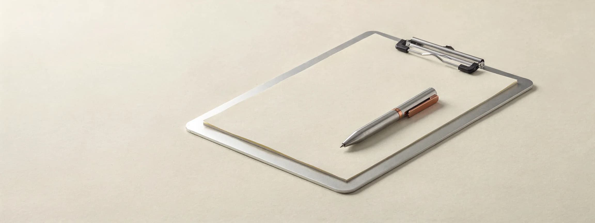 A machined aluminium clipboard holding a sheet of paper, a steel pen with a copper clip across it.