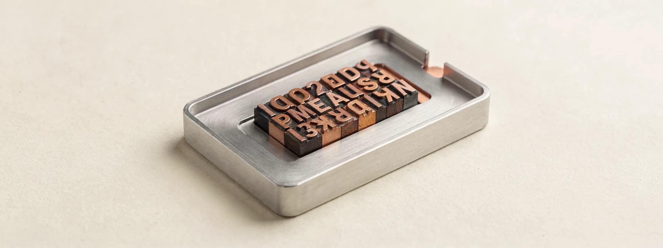 A machined steel type tray holding a few copper letterpress sorts, on paper.